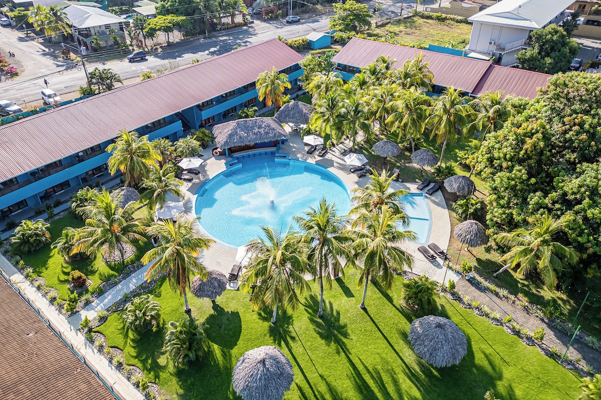 An aerial view of a resort with a central swimming pool surrounded by palm trees and thatched umbrellas, with buildings in the background.