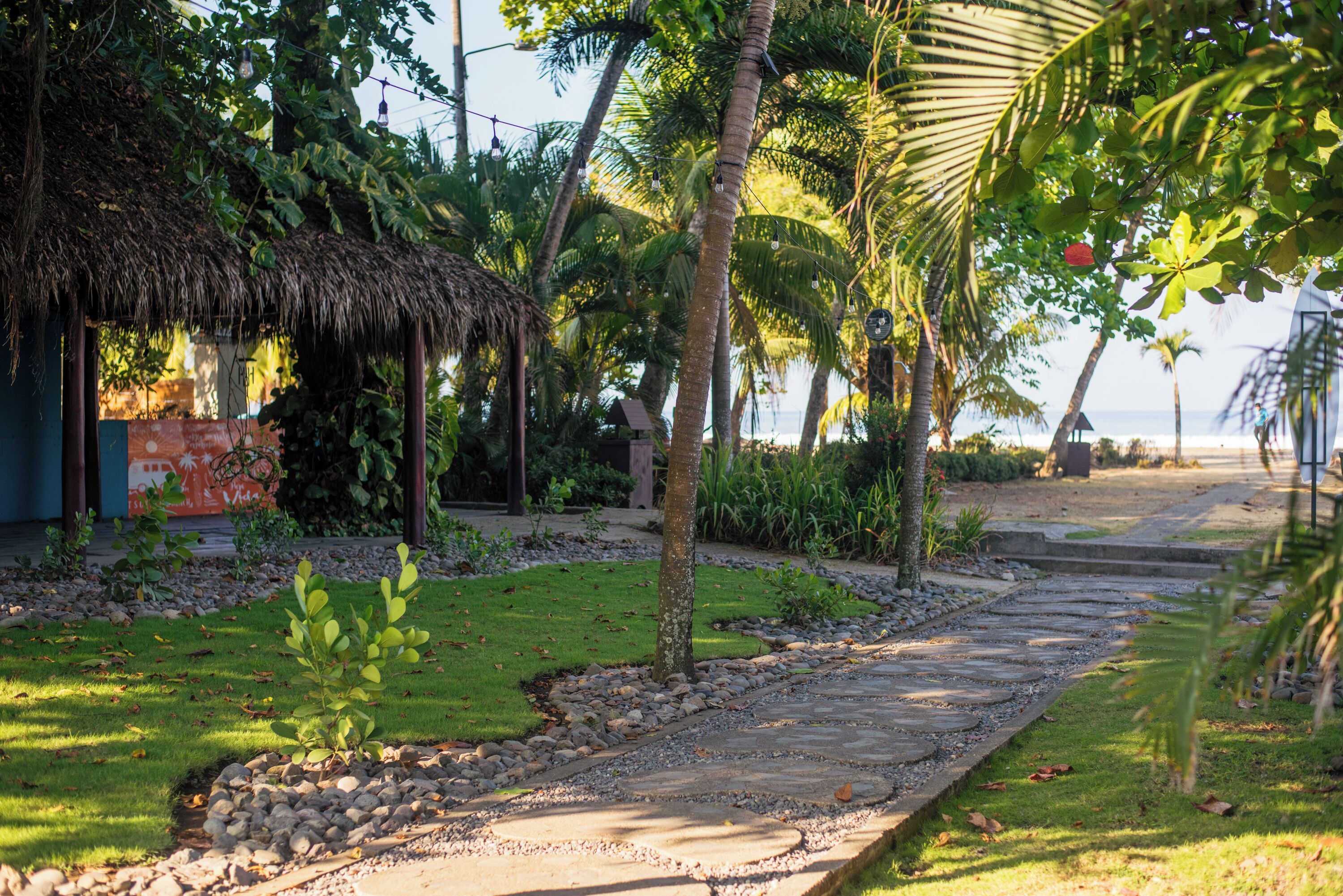 Un jardín tropical con una cabaña de techo de paja, palmeras y un sendero de piedra que lleva a una playa.