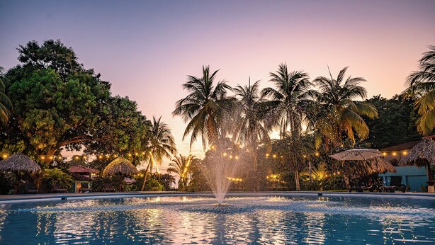 A swimming pool with a water fountain, surrounded by palm trees and thatched umbrellas, under a twilight sky. - Beach Break Resort (Jacó)