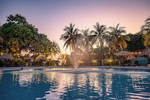 A swimming pool with a water fountain, surrounded by palm trees and thatched umbrellas, under a twilight sky. - Beach Break Resort (Jacó)