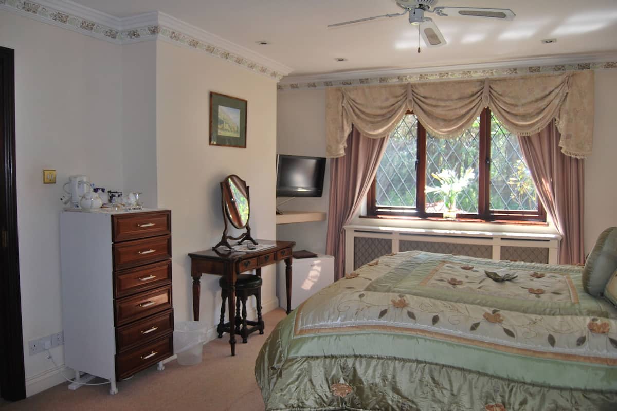 A bedroom with a bed, dresser, vanity, television, and a window with curtains.