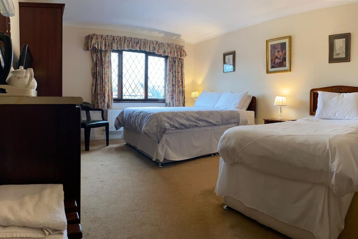 A hotel room with two beds, a desk with a kettle and teapot, a chair, and a window with curtains.