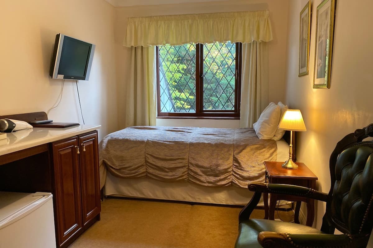 A bedroom with a bed, a desk with a computer, a chair, a lamp, and a window with a view of trees.