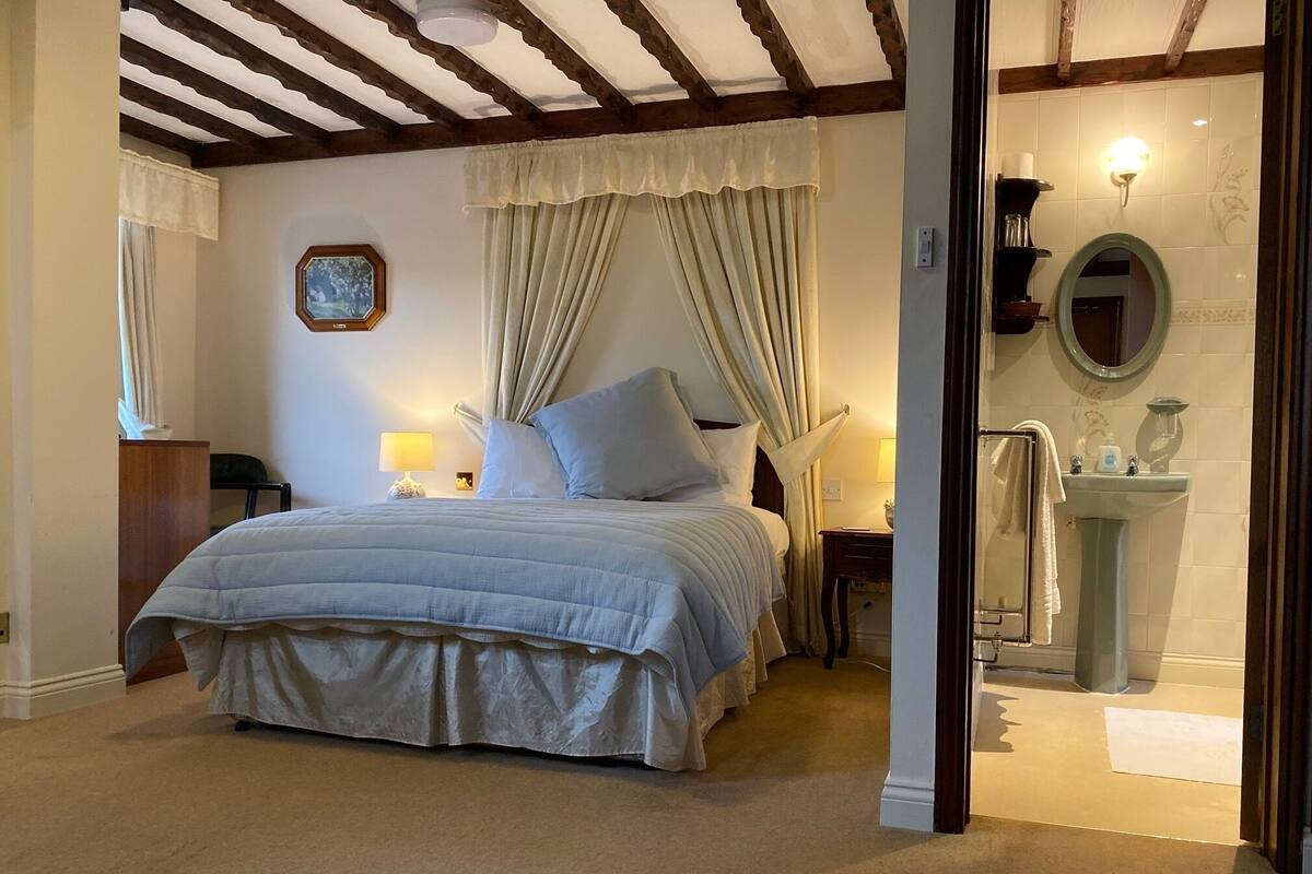 A bedroom with a bed, a nightstand, a lamp, a window with curtains, and a view into the bathroom.