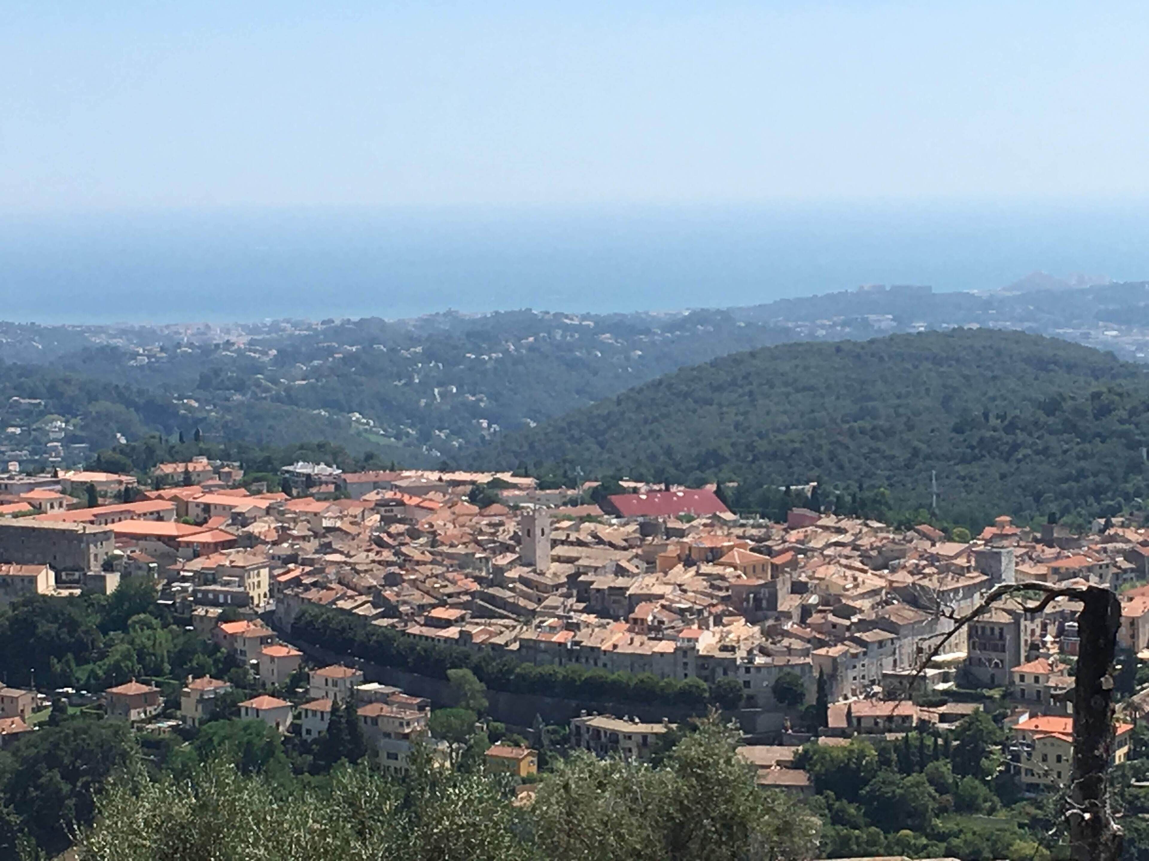 A panoramic view of a historic town with dense red-roofed buildings, a prominent tower, and lush green hills in the background.
