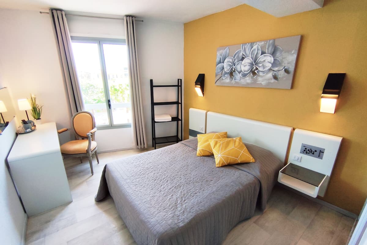 A hotel room with a bed, a desk, a chair, a window with curtains, a painting on the wall, and a bedside table with a control panel.