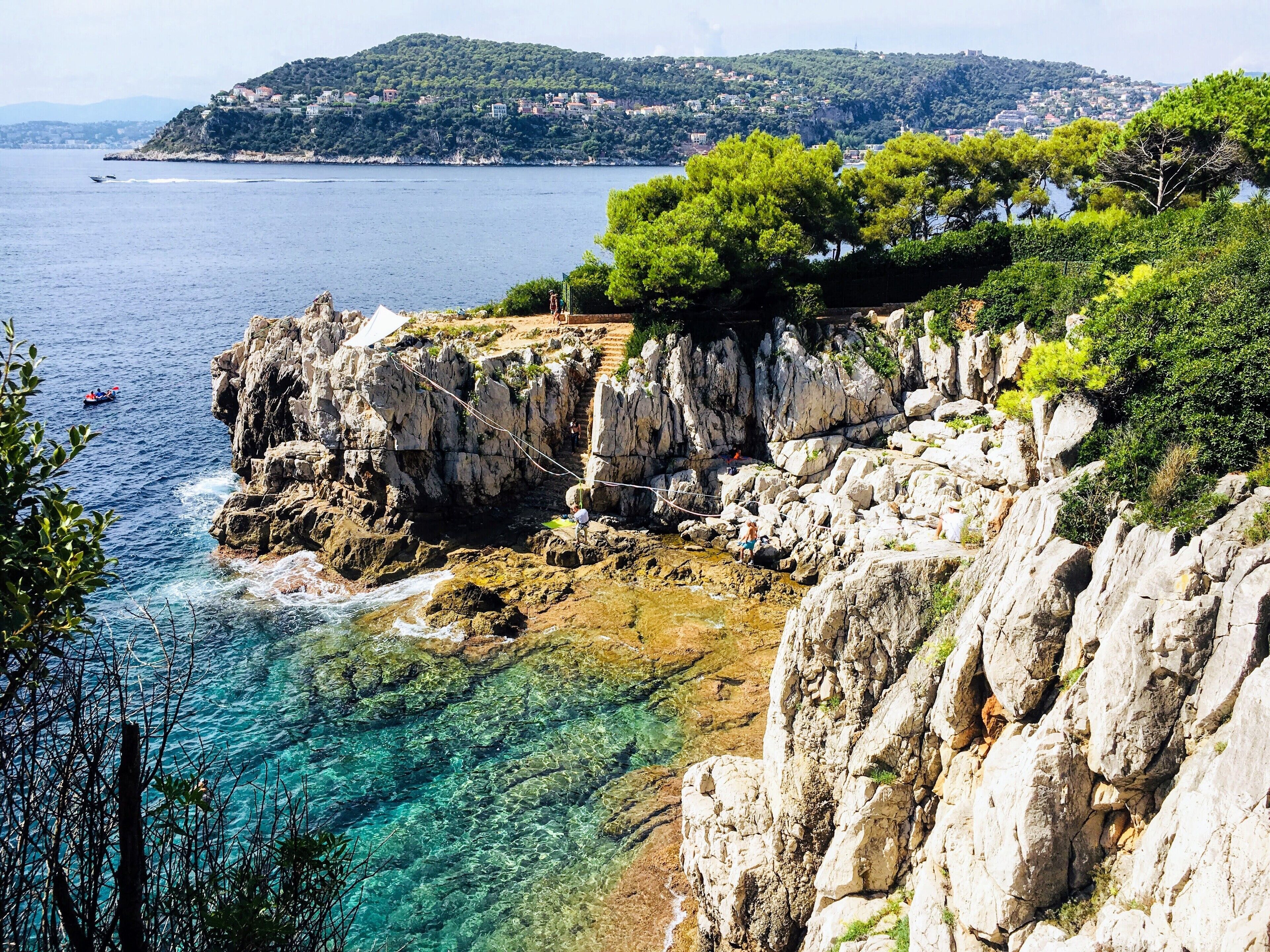 A coastal landscape with rocky cliffs, clear blue waters, and a distant view of a town on a hill.