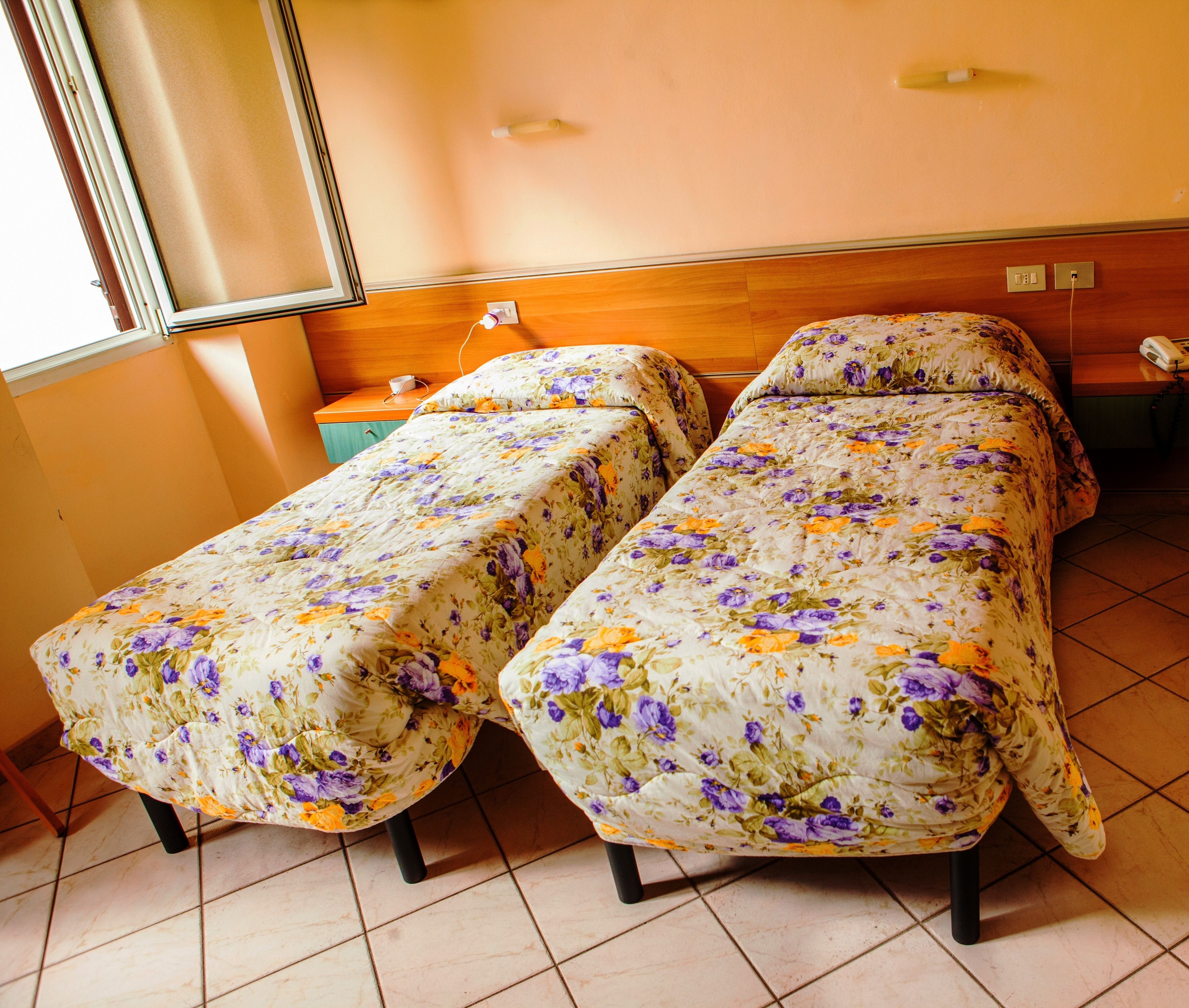 Habitación con dos camas cubiertas con ropa de cama estampada, un cabecero de madera, una mesita de noche con una lámpara y una ventana con persianas.
