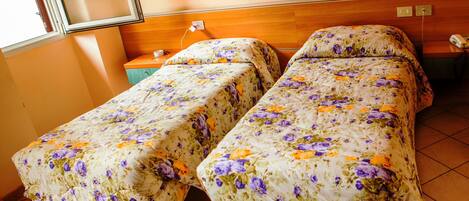 Quarto com duas camas cobertas por roupa de cama floral, cabeceira de madeira, mesa de cabeceira com abajur e janela com persianas.