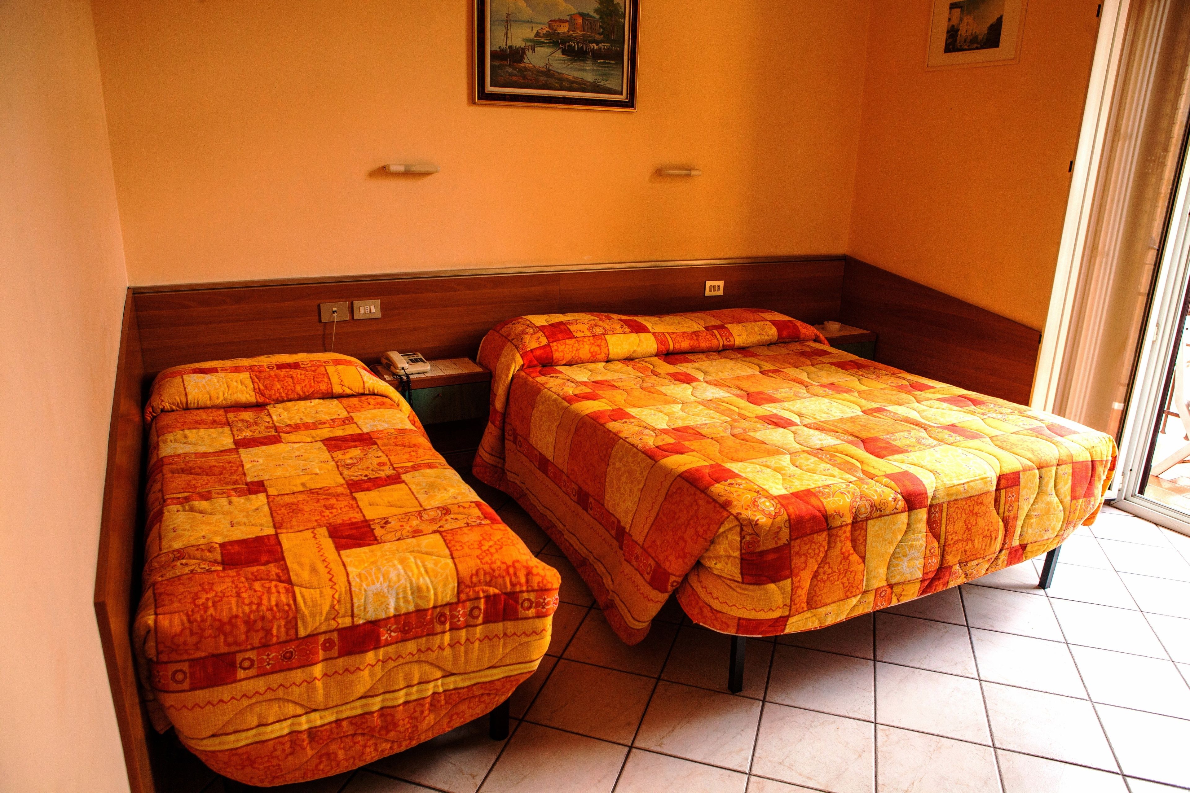A hotel room with two single beds, a wooden headboard, a painting on the wall, and a tiled floor.