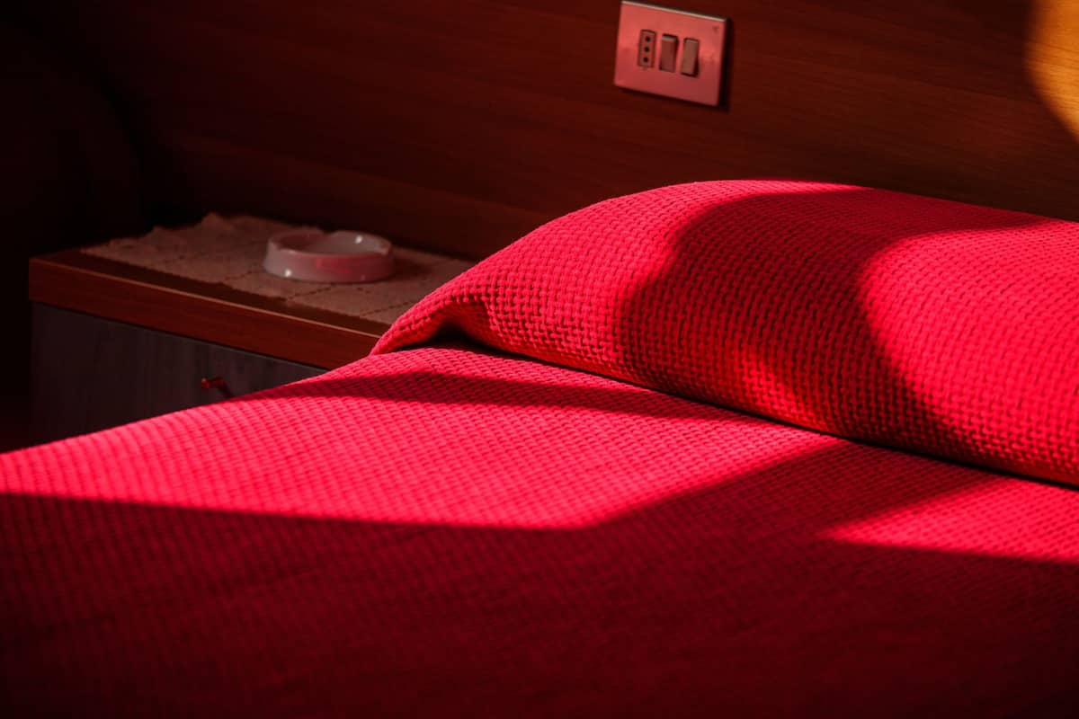 Un lit avec une couverture rouge, une tête de lit en bois et une table de chevet sur laquelle se trouvent une lampe et un bol.