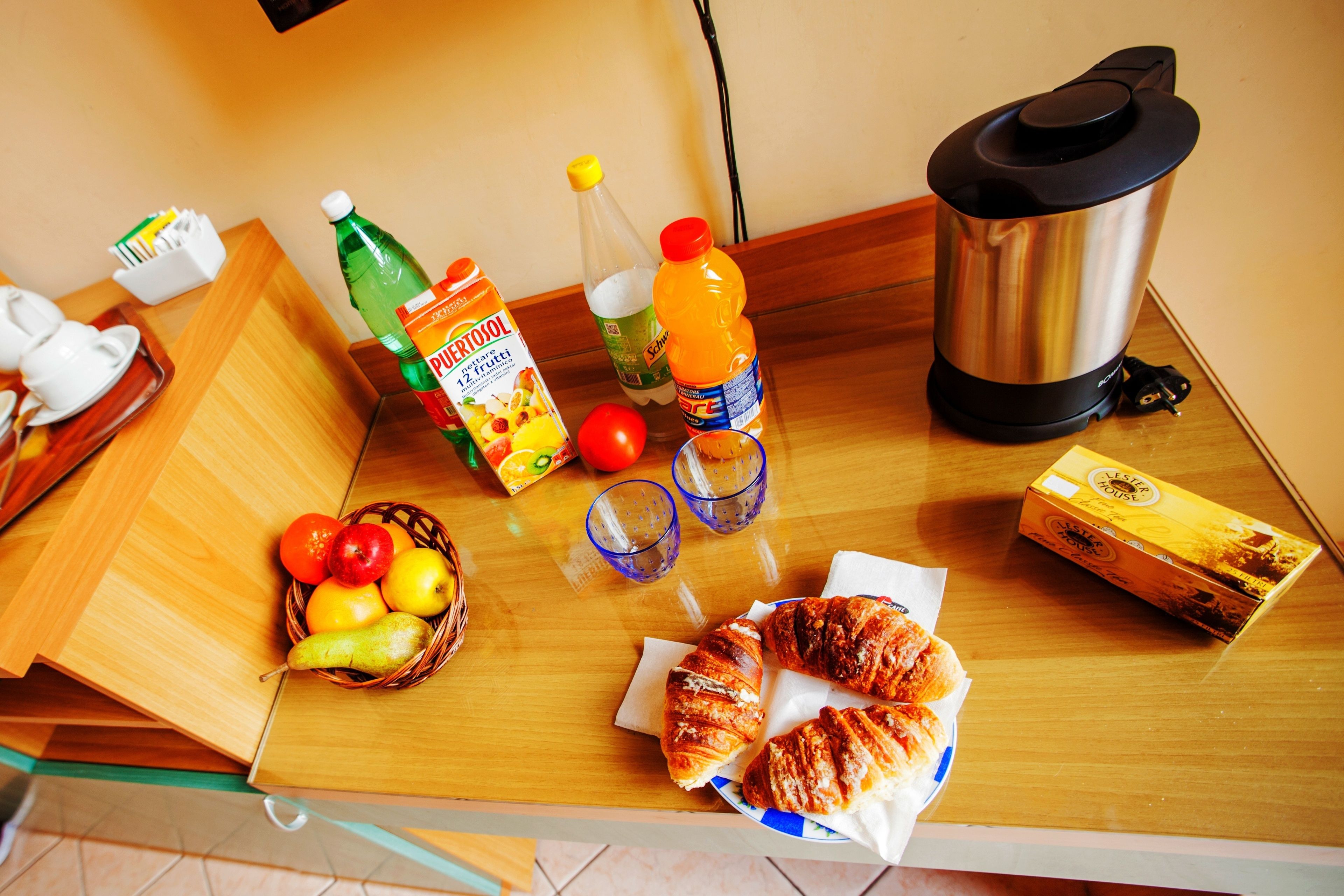 A breakfast table with croissants, a fruit basket, juice, and a coffee maker.