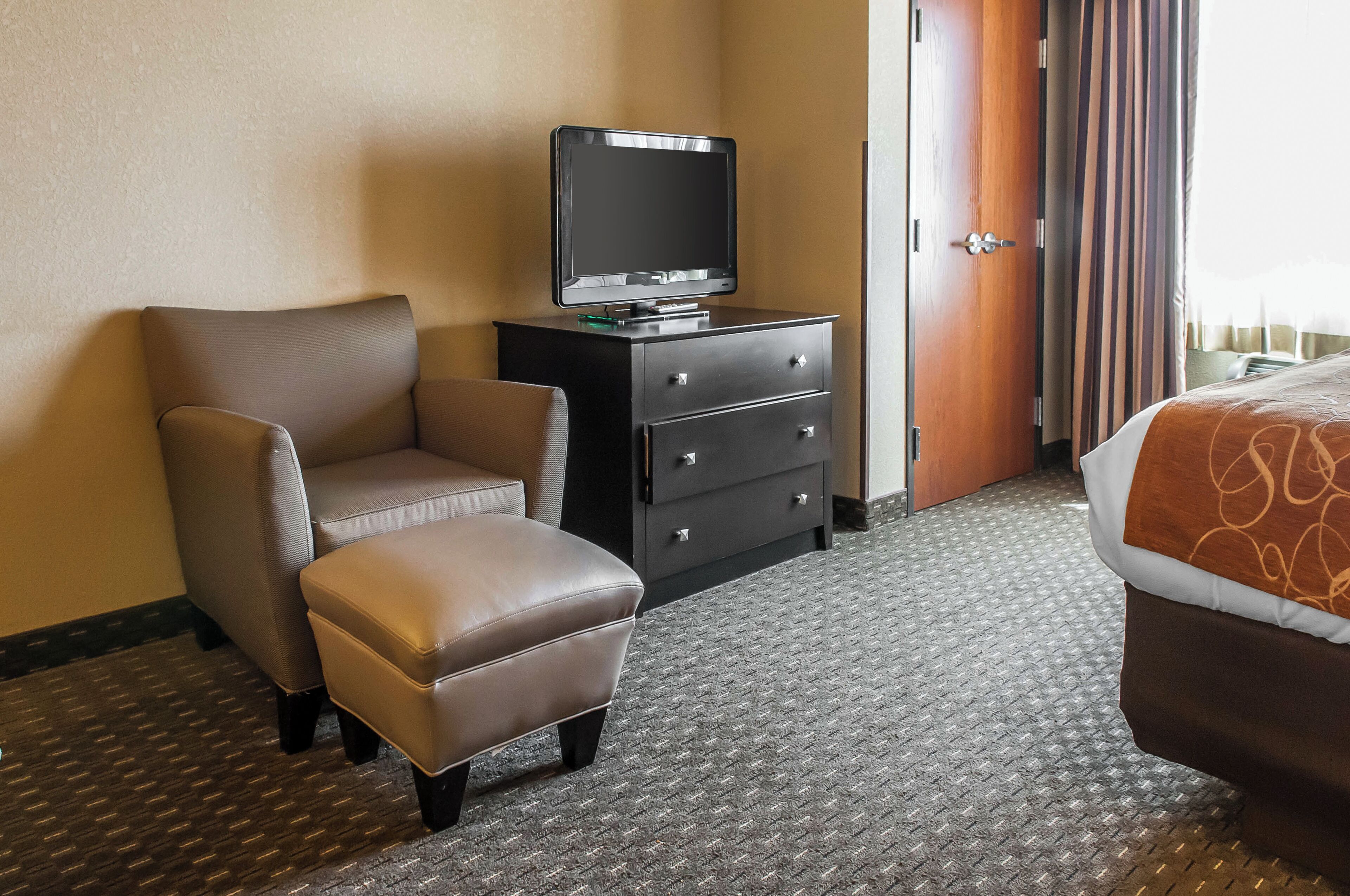 Une chambre d’hôtel avec un fauteuil en cuir marron, un petit pouf, une commode noire sur laquelle est posée une télévision, et un lit recouvert d’une couvre-lit à motifs.