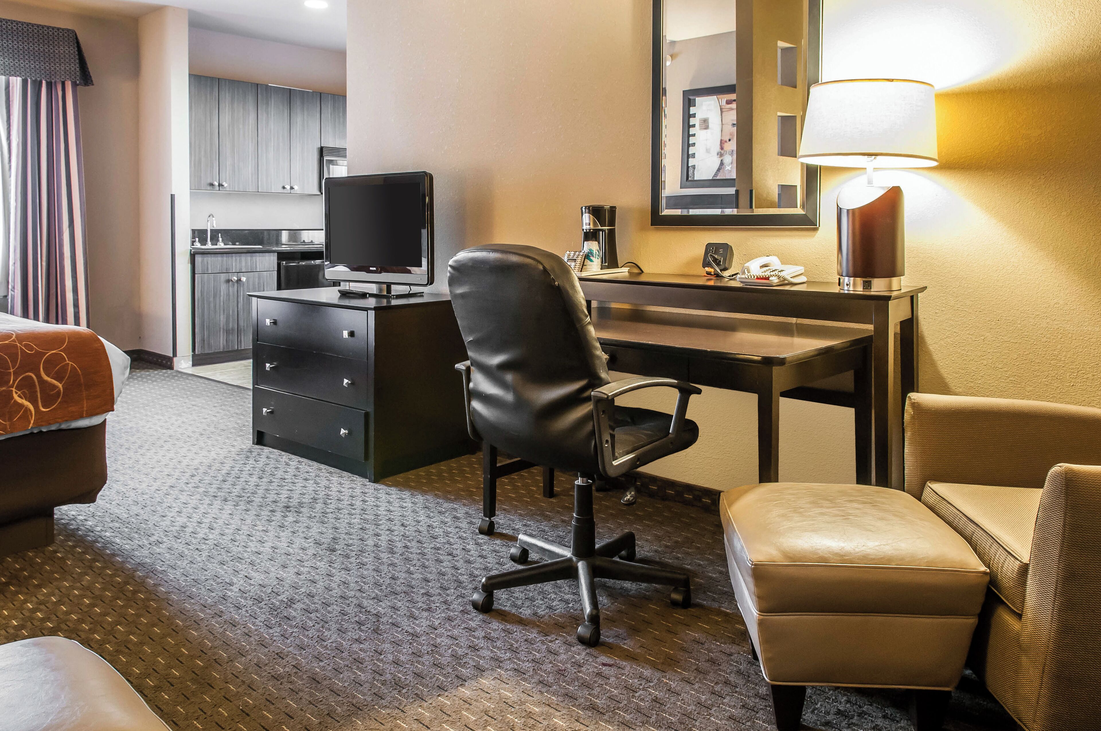 A hotel room with a desk, chair, and a bed. There is a kitchenette in the background.