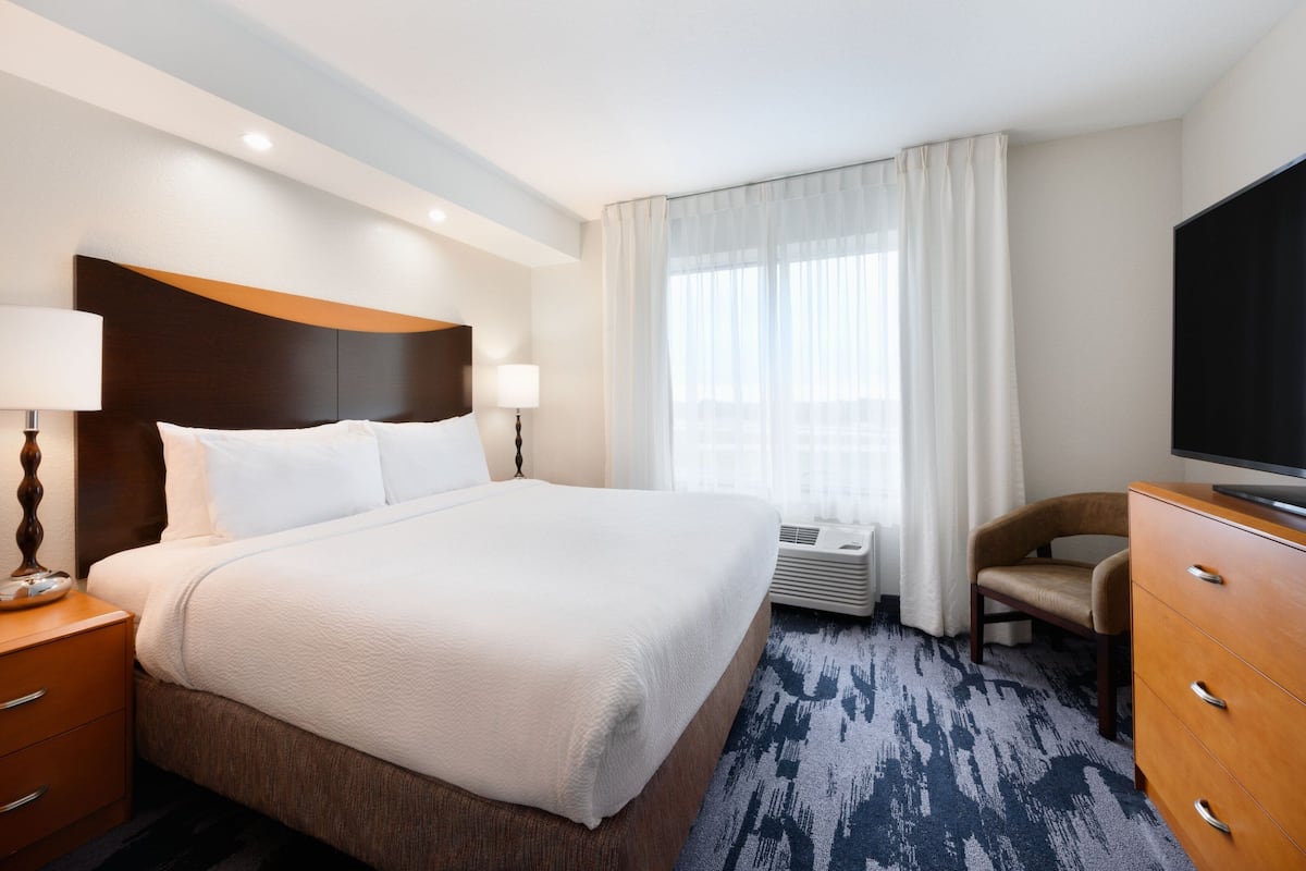 A hotel room with a large bed, a desk with a lamp, a chair, a TV, and a window with curtains.
