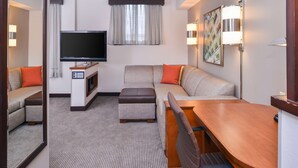 Habitación de hotel moderna con sofá, reposapiés y una mesa de madera. Un televisor de pantalla plana está montado en la pared, y hay dos lámparas de pared.