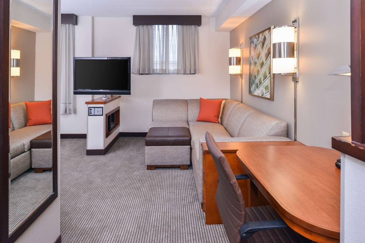 Ein modernes Hotelzimmer mit einer Couch, Hocker und einem Holztisch. Ein Flachbildfernseher ist an der Wand befestigt, und es gibt zwei Wandleuchten.