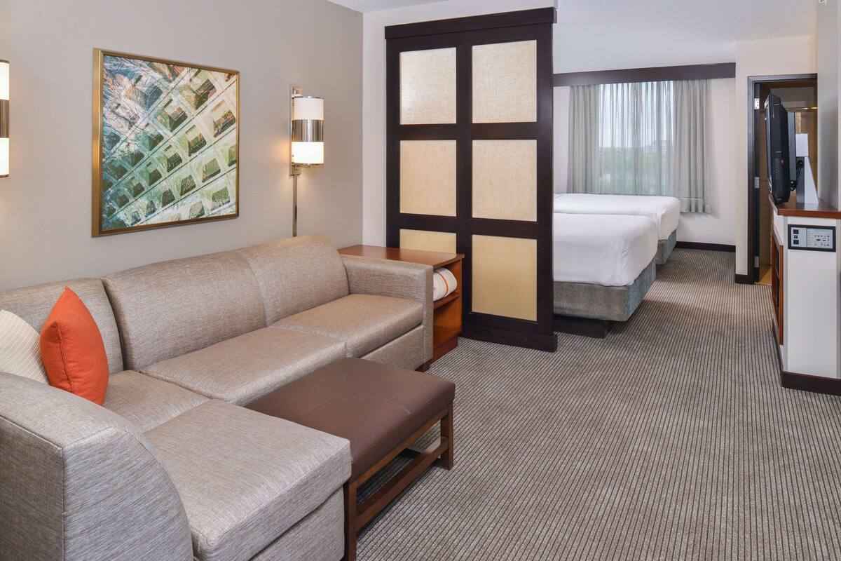 Ein Hotelzimmer mit einer Couch, einem kleinen Tisch, einem Schreibtisch mit Computer sowie einem Bett und einem großen Fenster mit Vorhängen.