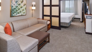 Habitación de hotel con sofá, una mesita, escritorio con computadora, cama y un ventanal grande con cortinas.