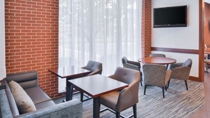 Habitación de hotel con pared de ladrillo, un ventanal con cortinas translúcidas, un televisor de pantalla plana montado en la pared, una mesa redonda con cuatro sillas y un sofá con cojines.