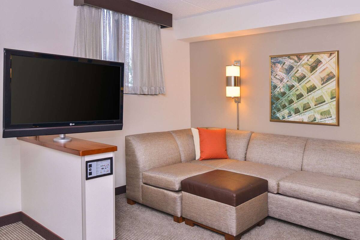 Ein modernes Hotelzimmer mit Flachbildfernseher, einem beigefarbenen Sofa mit braunem Hocker, zwei Wandlampen und einem gerahmten abstrakten Gemälde.