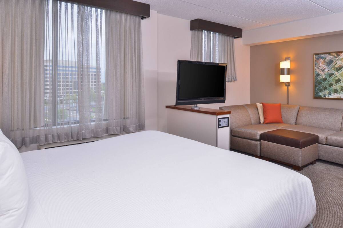 Ein Hotelzimmer mit einem großen Bett, einem Fernseher auf einem Schrank, einer Couch mit Hocker und einem Bild an der Wand.