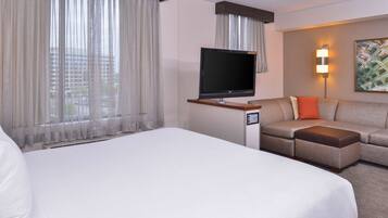 A hotel room with a large bed, a TV on a stand, a sofa with an ottoman, and a painting on the wall.