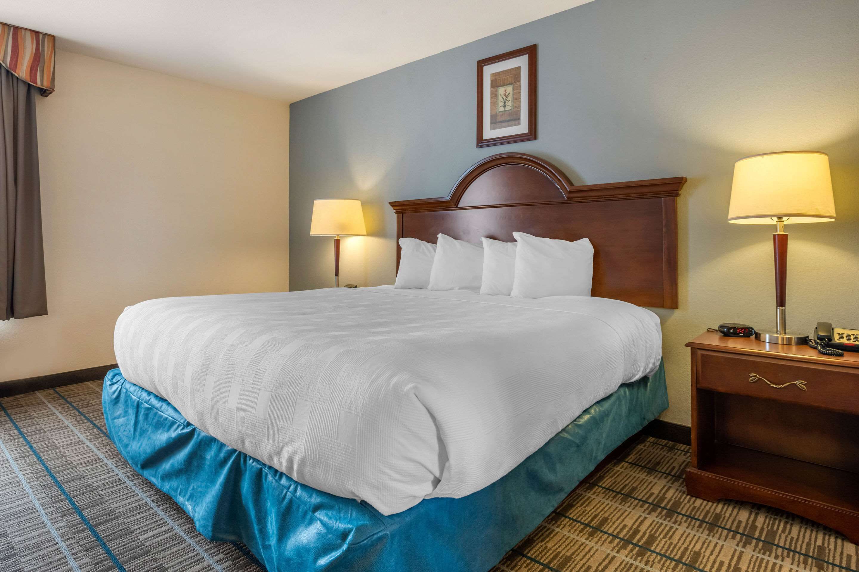Una habitación de hotel con una cama grande, dos lámparas de noche, una mesita de noche de madera y un cuadro enmarcado en la pared.