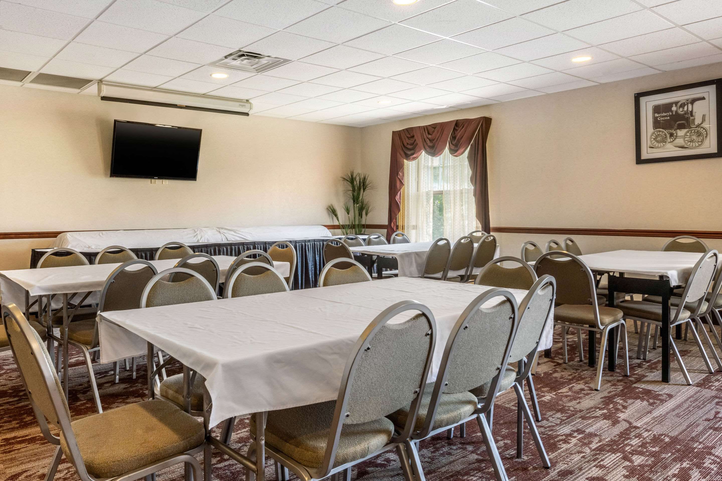 Une salle de conférence équipée de plusieurs tables et chaises, d’un téléviseur à écran plat fixé au mur et d’une fenêtre avec des rideaux.