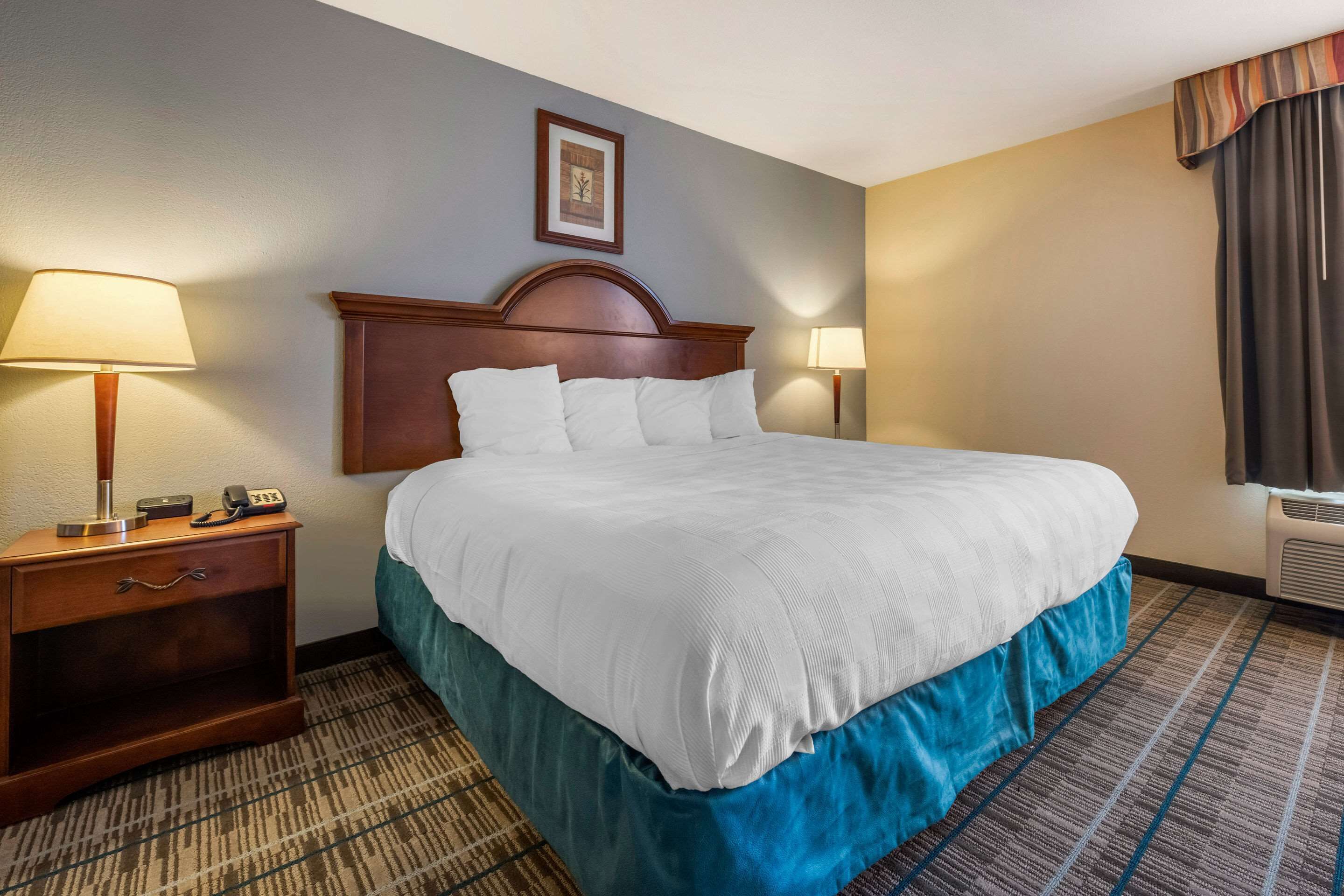 Un cuarto de hotel con una cama grande, una mesita de noche de madera con una lámpara, un cuadro en la pared y una ventana con cortinas.