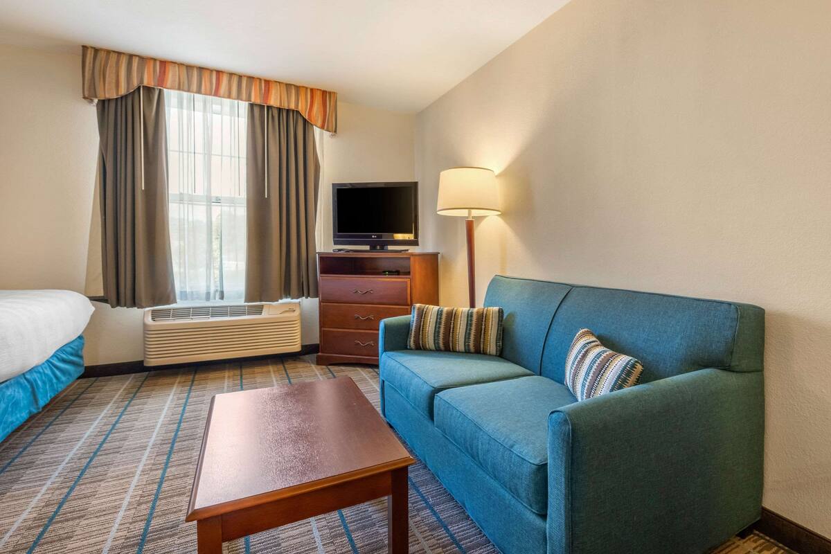 Ein Hotelzimmer mit einem Bett, einem blauen Sofa, einem Holztisch, einem Fernseher, einer Lampe und einem Fenster mit Vorhängen.