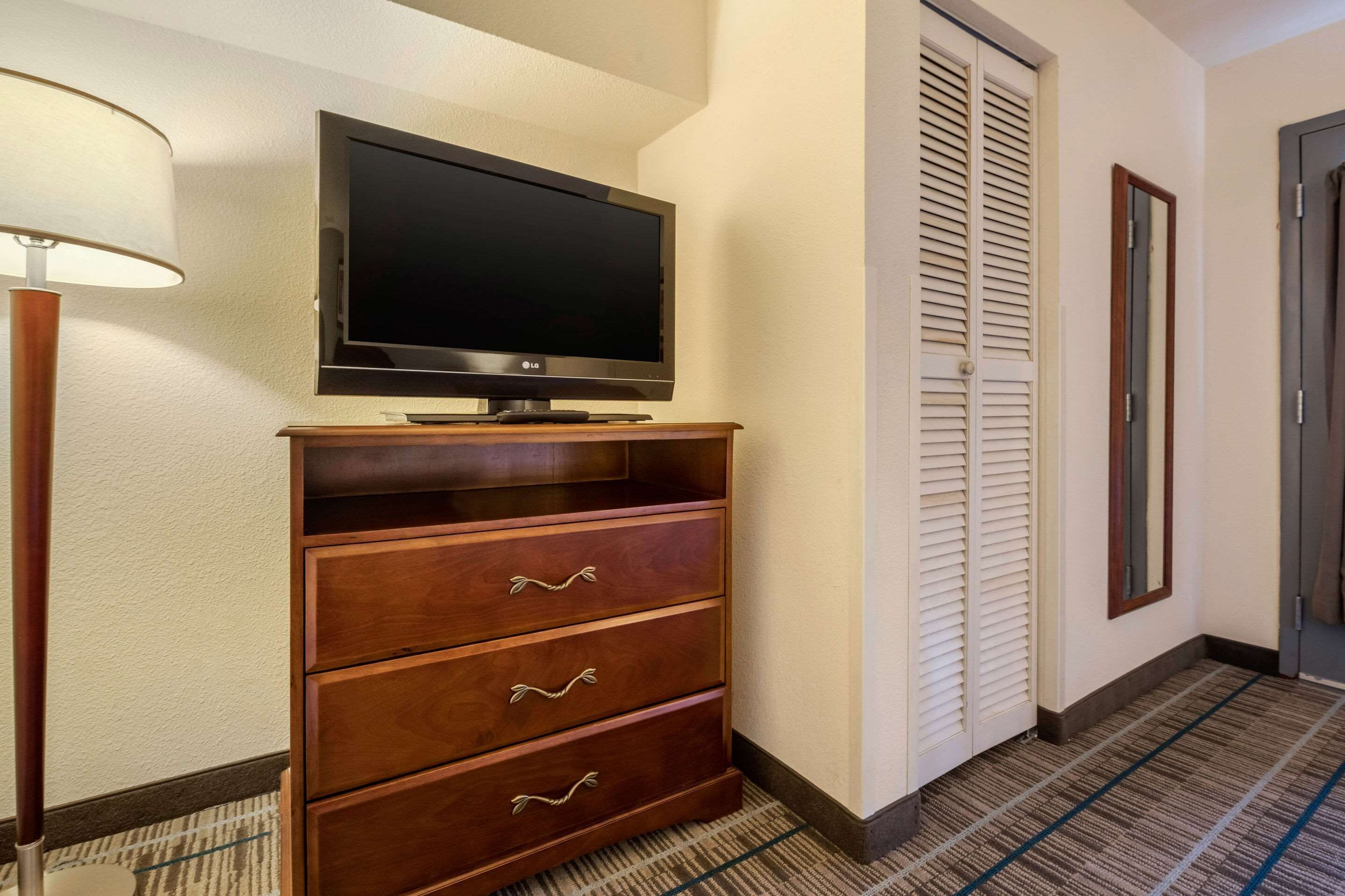 A hotel room with a flat-screen TV on a wooden cabinet, a lamp, a closet with white louvered doors, and a patterned carpet.