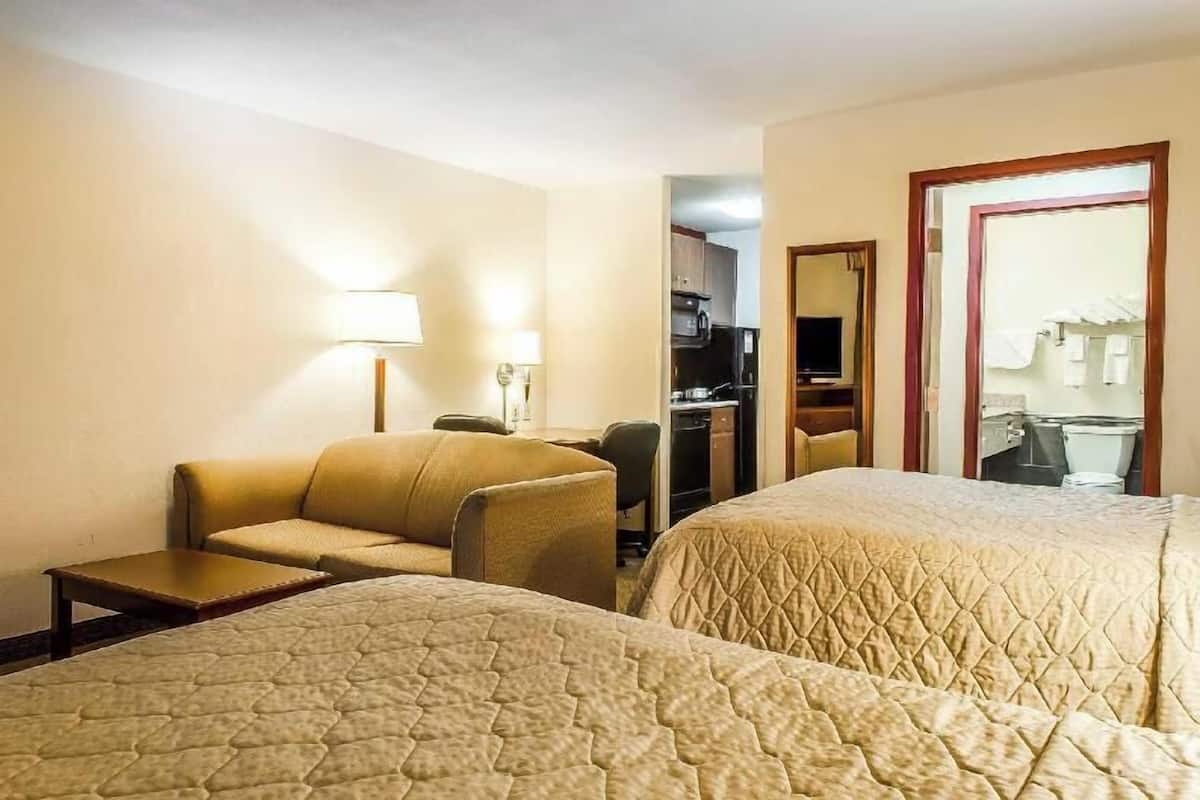 Ein Hotelzimmer mit zwei Betten, einem Sofa, einem Schreibtisch, einer Lampe und Blick auf den Küchenbereich.