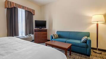 A hotel room with a large bed, a blue sofa, a wooden nightstand, a flat-screen TV, a lamp, and a window with curtains.