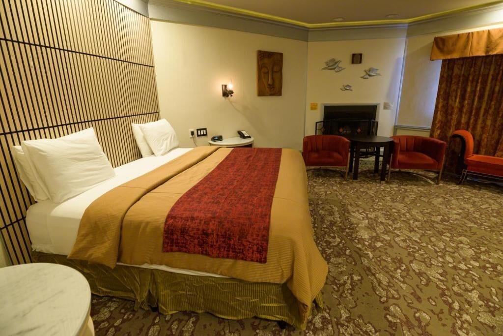 A hotel room with a bed, a small table, two chairs, a wall-mounted light, and a fireplace.