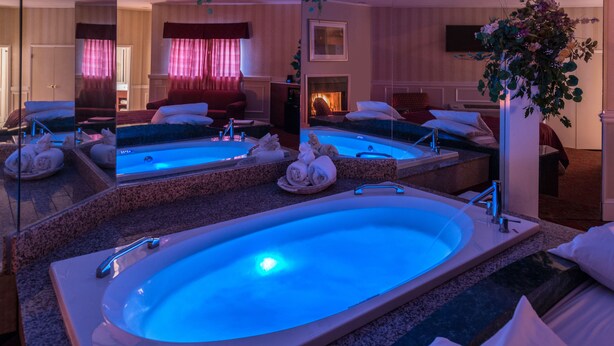 A hotel room with a large oval bathtub, a fireplace, a bed, and a mirror reflecting the room. - Inn of the Dove Romantic Suites with Jetted Tub & Fireplace (Bensalem)