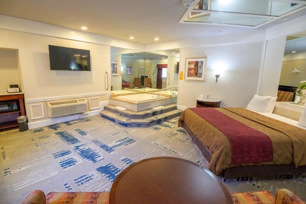 Habitación de hotel con una cama grande, una zona de estar con una mesa, un espejo y un televisor montado en la pared.