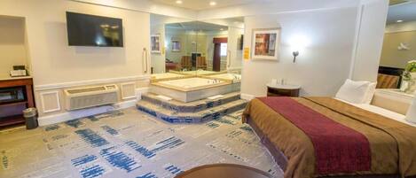Habitación de hotel con una cama grande, una zona de estar con una mesa, un espejo y un televisor montado en la pared.
