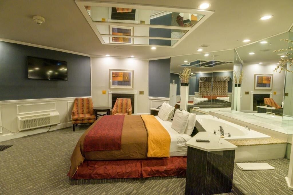 A hotel room with a bed, a desk, a large mirror, a television, and a bathroom with a bathtub and sink.