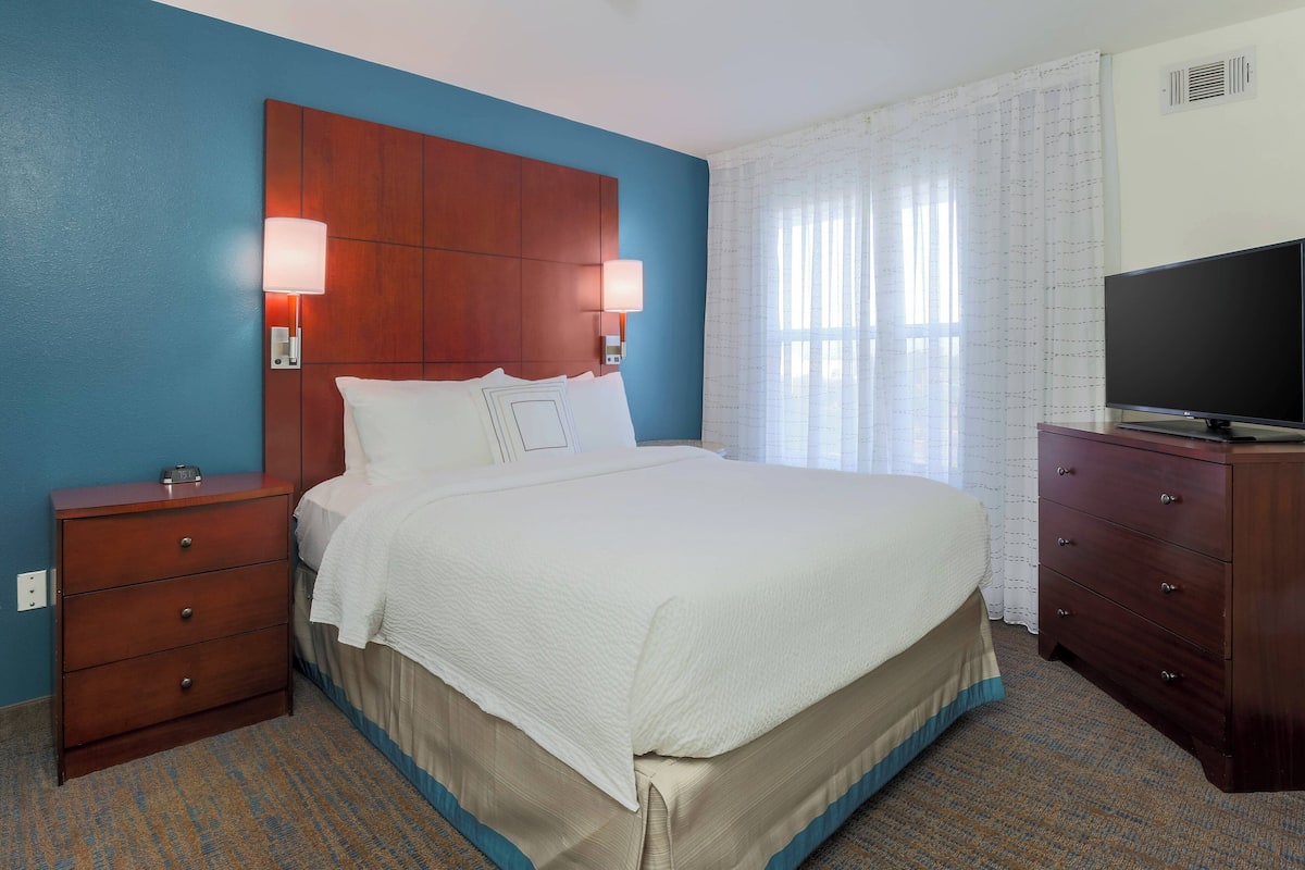 A hotel room with a large bed, a wooden nightstand, a dresser, a television, and a window with curtains.