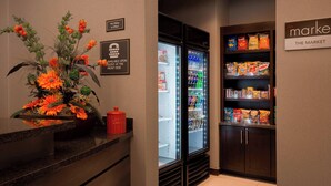 A hotel reception area with a flower arrangement, a pet policy sign, a refrigerator stocked with beverages, and a snack shelf. - Residence Inn by Marriott Dallas Arlington South (Arlington)