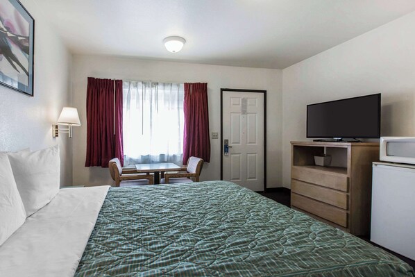 A hotel room with a bed, a desk with a chair, a television, a dresser, and a door.