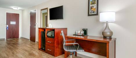 A hotel room with a wooden desk, a modern chair, a flat-screen TV, a coffee maker, and a lamp.