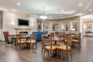 A hotel dining area with multiple tables and chairs, a flat-screen TV mounted on the wall, and a fireplace. - Comfort Inn & Suites Van Buren - Fort Smith (Van Buren)