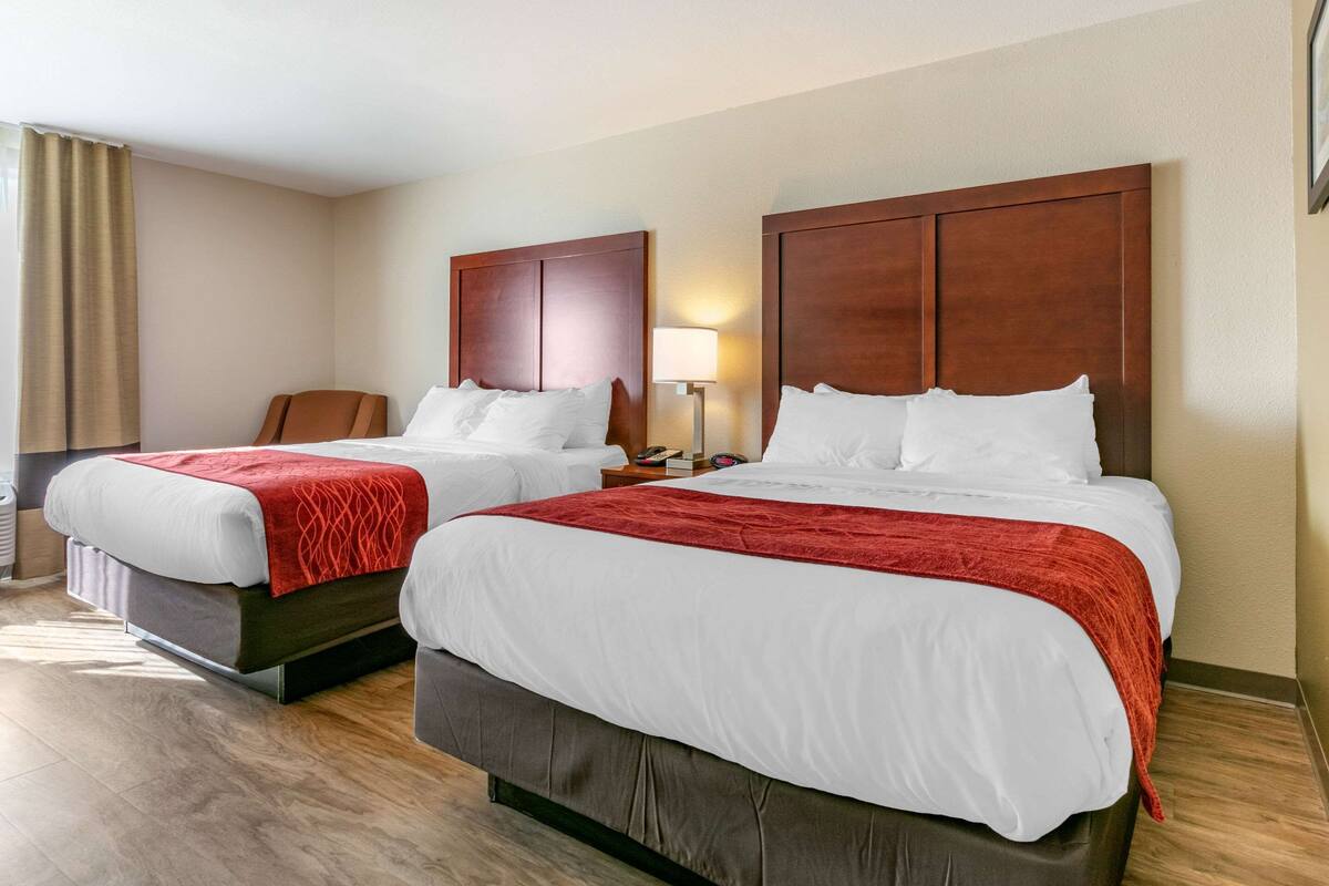 A hotel room with two beds, each with white bedding and a red blanket. There is a wooden headboard, a nightstand with a lamp, and a chair.