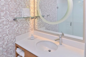 Un baño con pared de mosaicos, repisa de vidrio con toallas, espejo redondo y lavamanos con grifo.