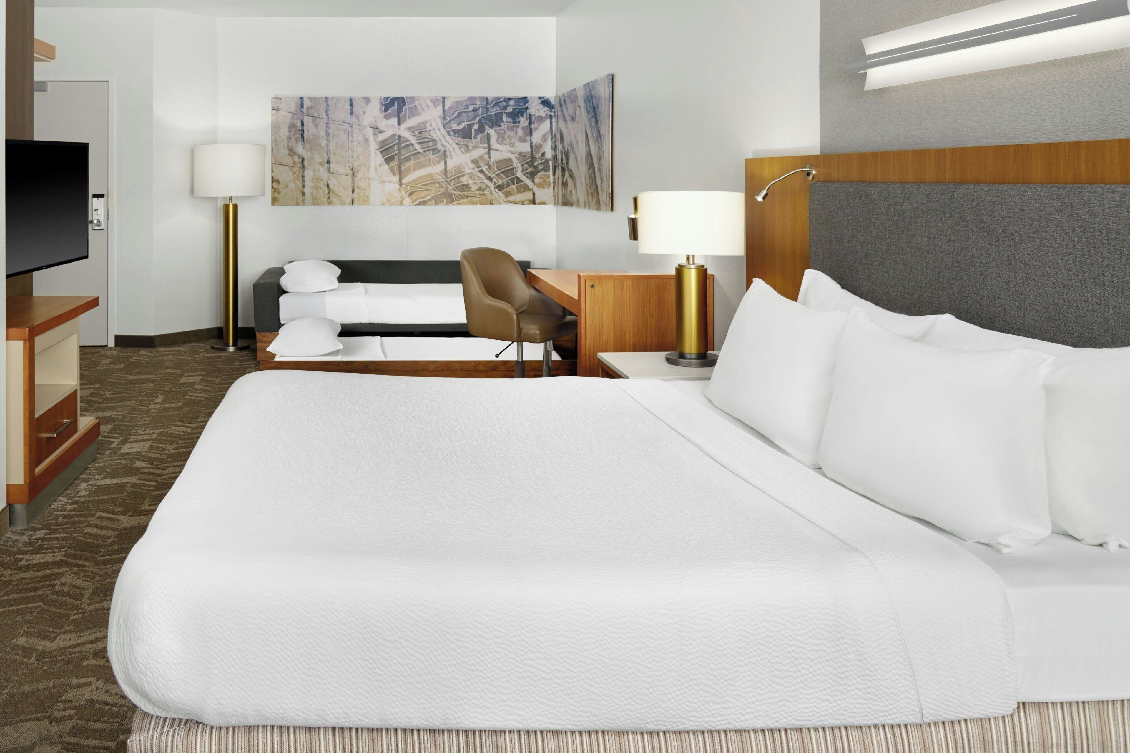 Una habitación de hotel con una cama grande, un escritorio, una silla, un televisor y un cuadro en la pared.