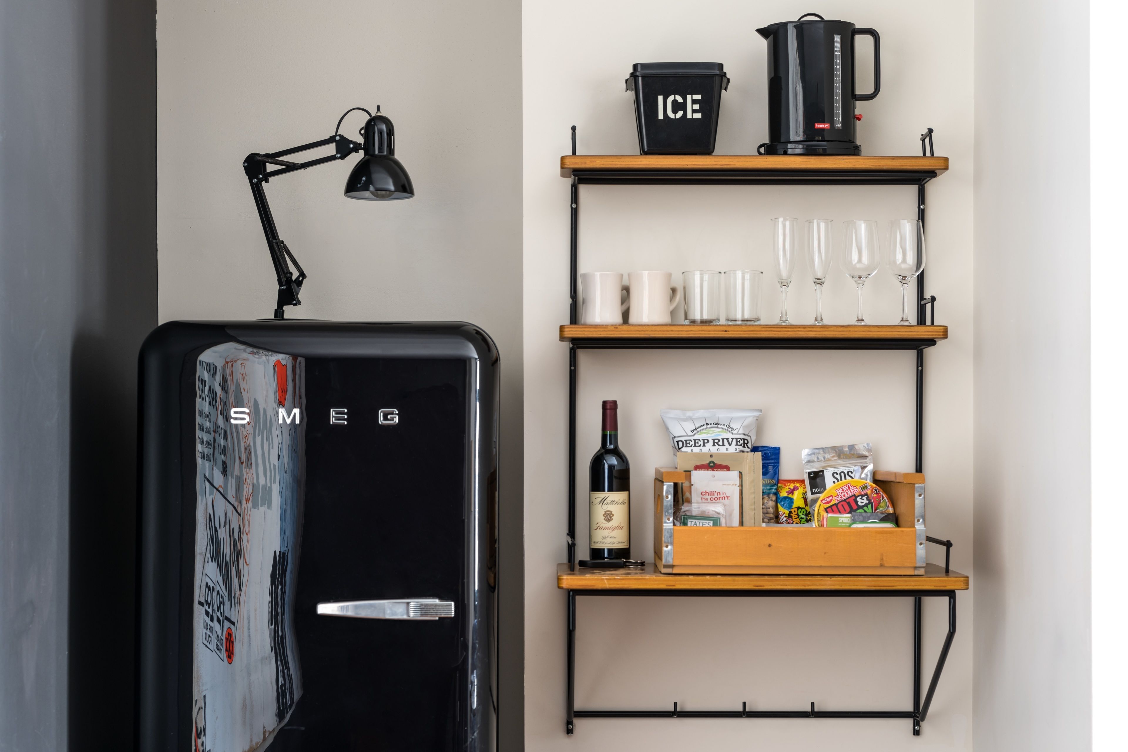 Un refrigerador compacto con compartimento congelador, una lámpara de escritorio negra y un estante de madera con una cafetera, vasos y aperitivos.
