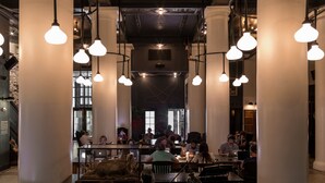 En rummelig café med høje lofter, synlige ventilationskanaler og hængende lamper. Bordene er indrettet til grupper, med et centralt siddeområde med lædersofaer.