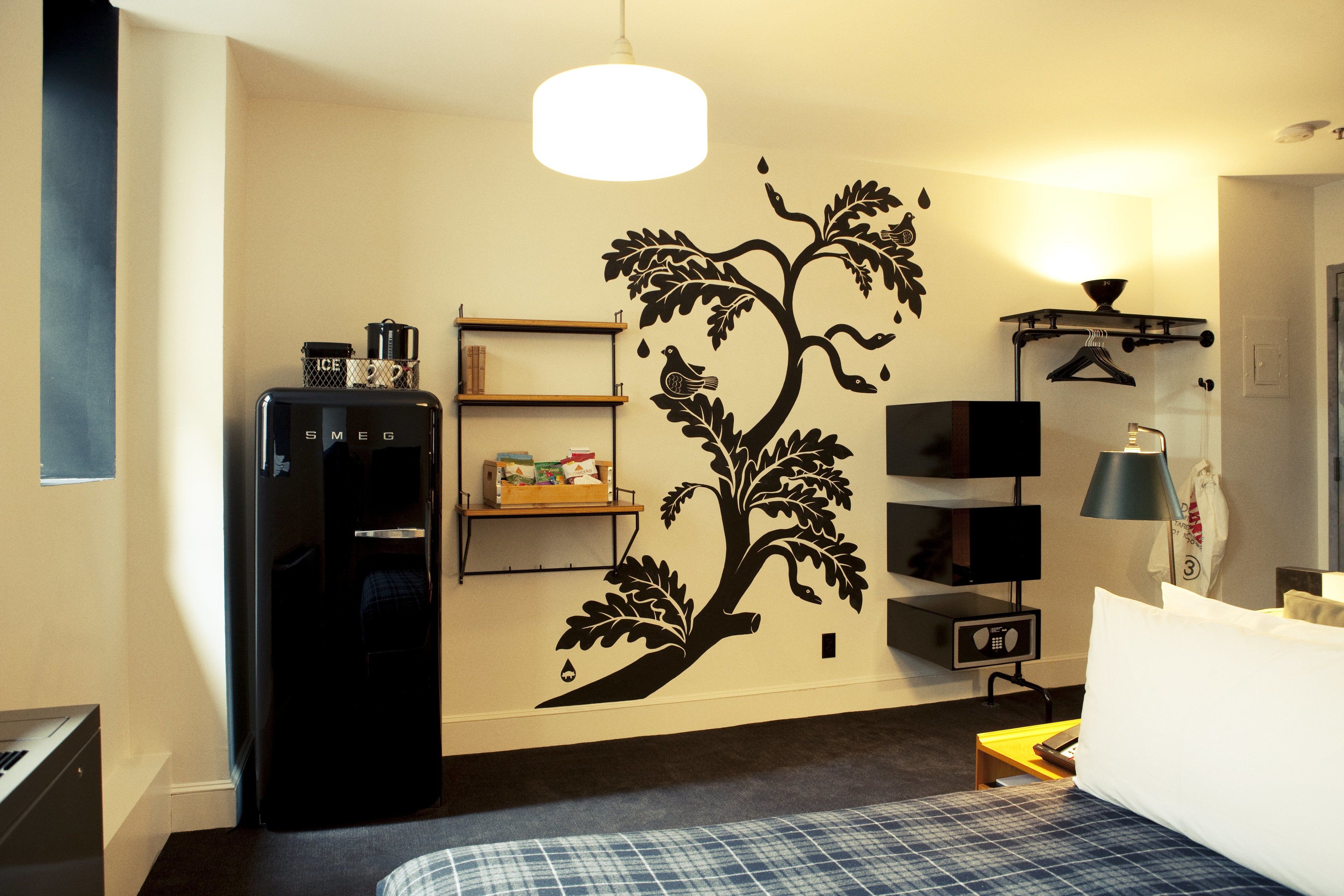 Une chambre d’hôtel avec un lit, un réfrigérateur noir, un téléviseur fixé au mur, une étagère avec des articles de toilette et un autocollant mural représentant un arbre.